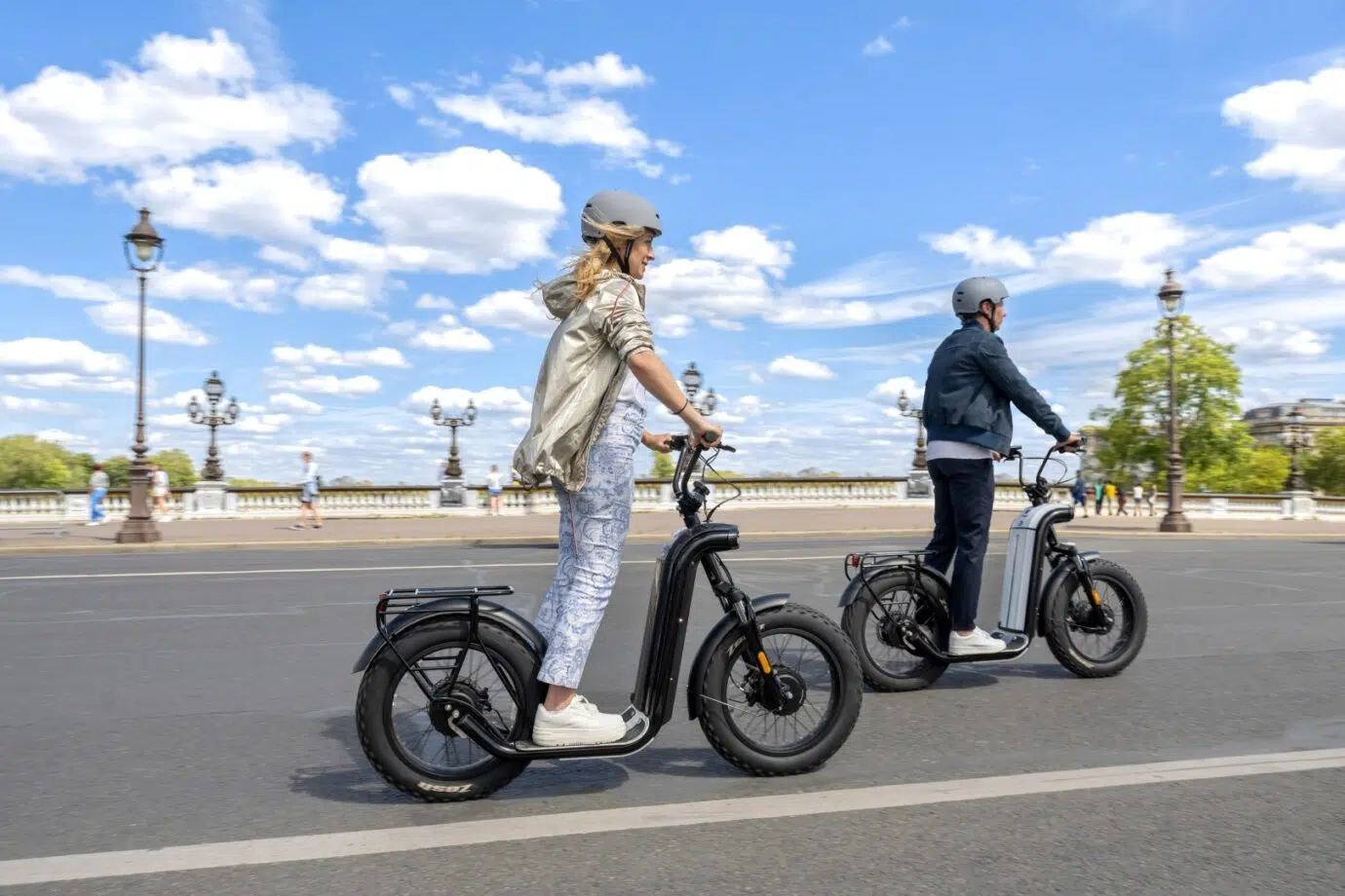 Deux personnes en trottinettes Zosh City traversant une place parisienne sous un ciel bleu